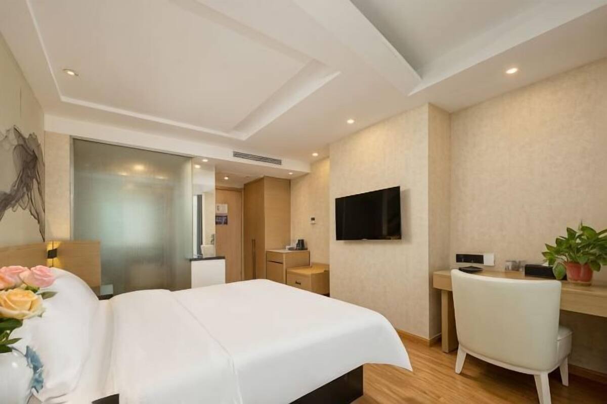 A hotel room with a bed, desk, chair, TV, and a potted plant.