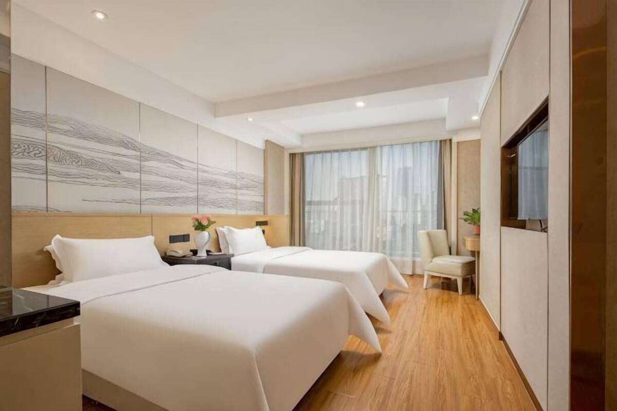 A hotel room with two beds, a desk, a chair, a TV, and a large window.