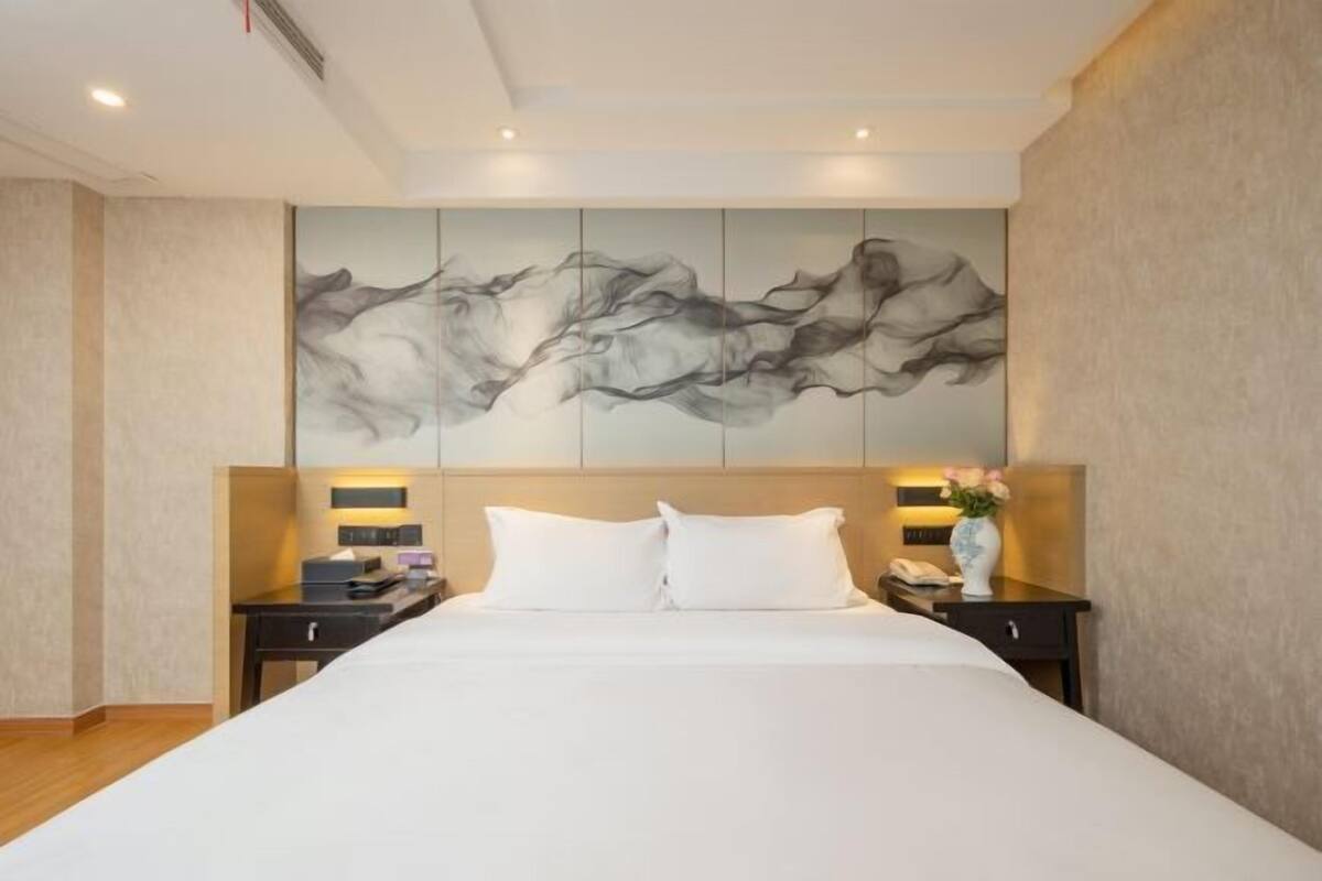 A hotel room with a large bed, two bedside tables, a nightstand, a wall-mounted artwork, and a vase with flowers.