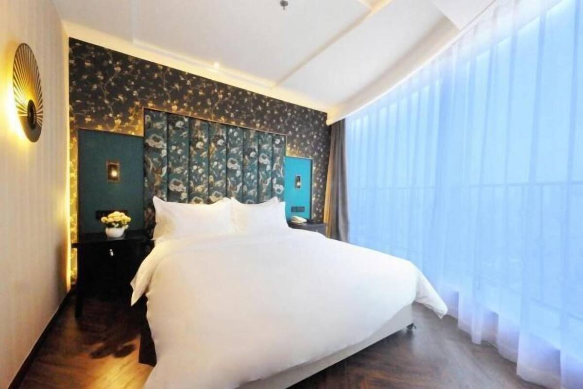 A hotel room with a large bed, a dark patterned headboard, a nightstand with a lamp, and a window with sheer curtains.