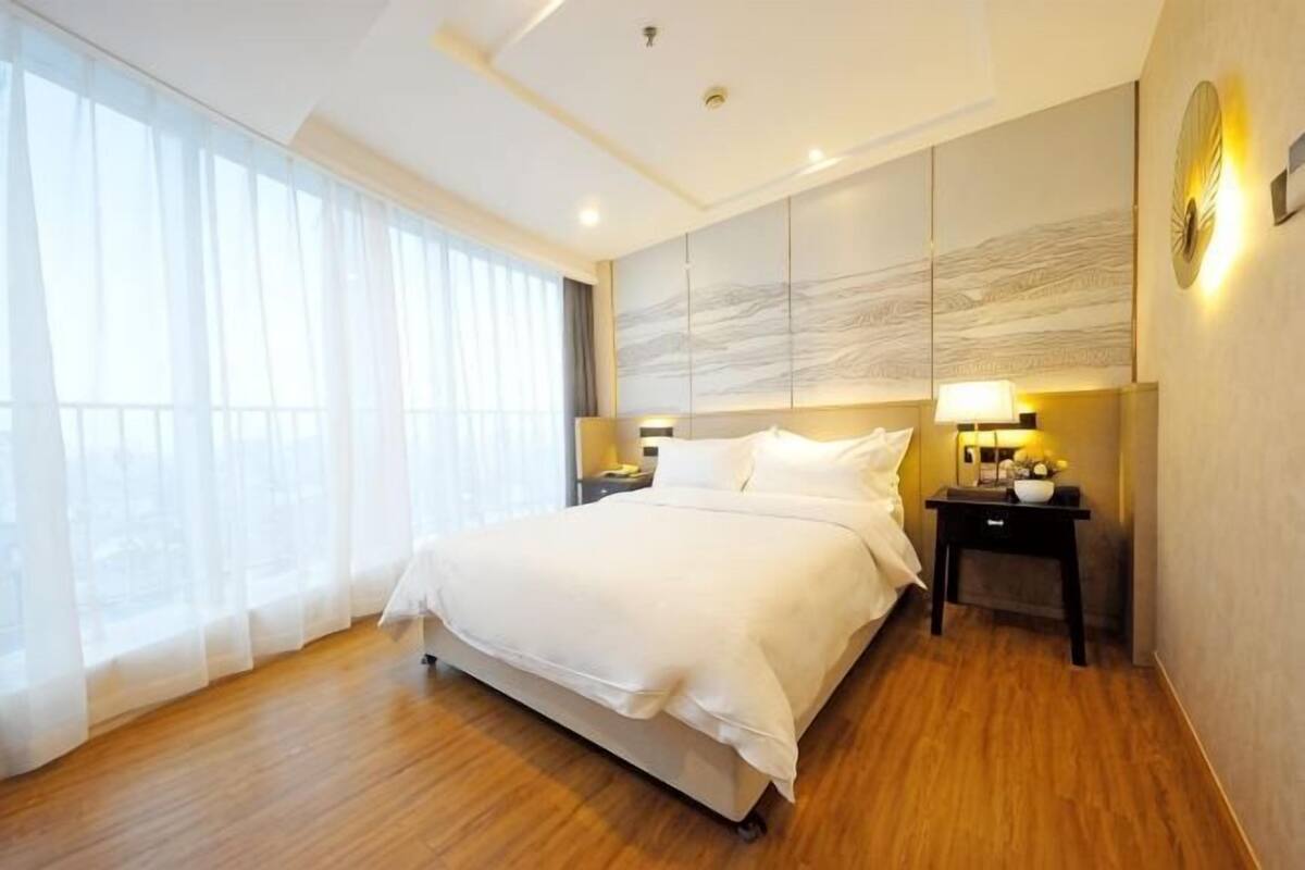 A hotel room with a large bed, a nightstand, a lamp, and a view through sheer curtains.