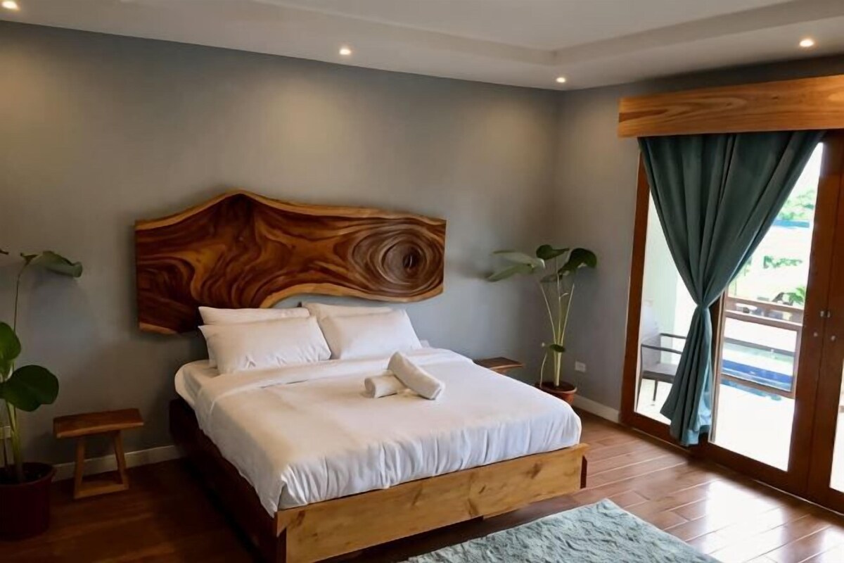 Un dormitorio con cabecera de madera, una cama con ropa blanca, dos mesitas de noche y un ventanal con cortinas.