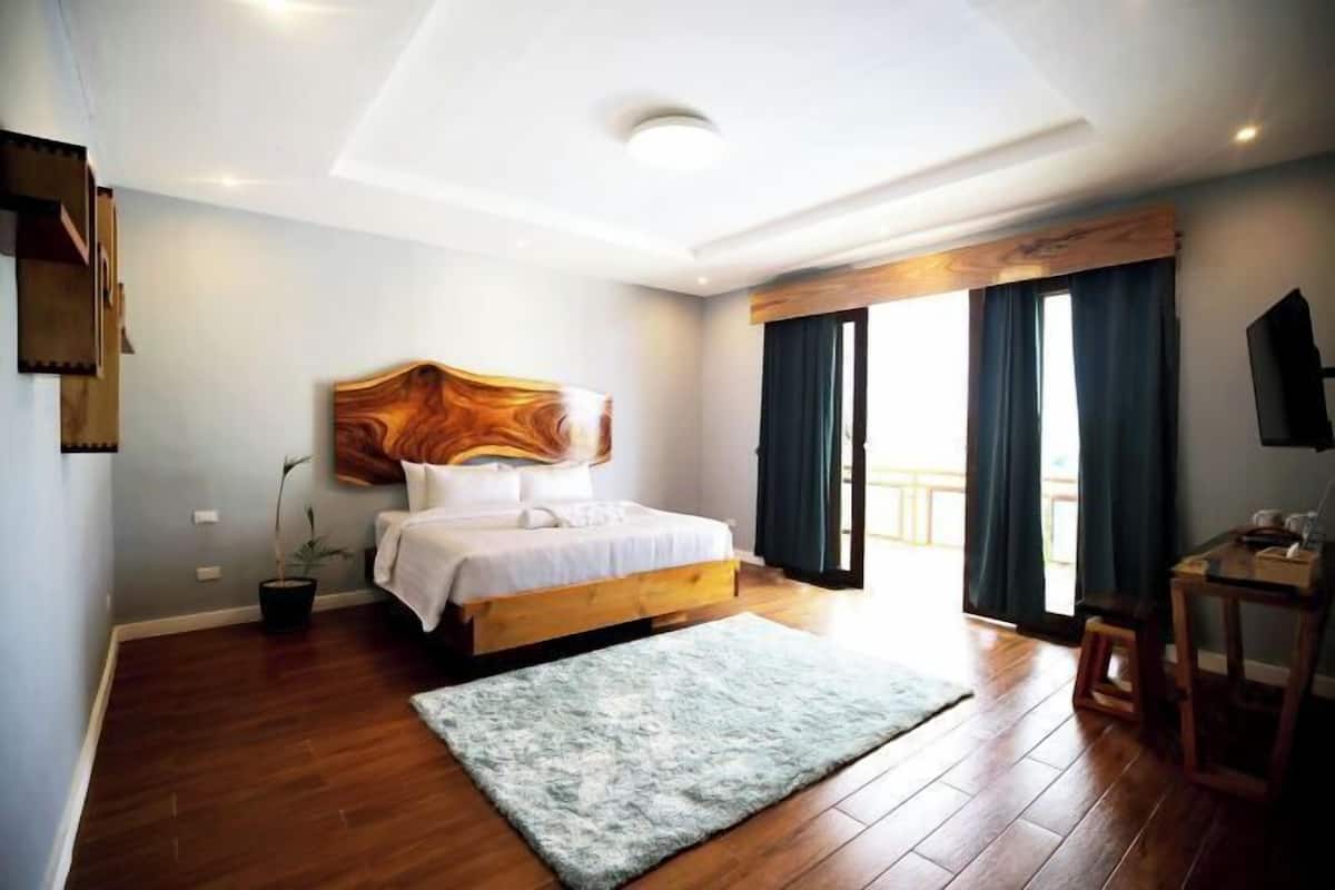 A bedroom with a wooden bed, a large window with curtains, a small table, and a potted plant.