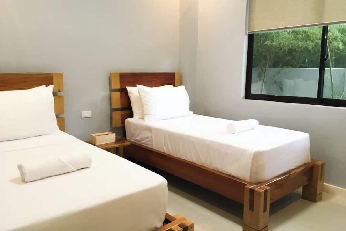A double bed with white linens, a wooden headboard, a bedside table, and a window with a view of greenery.