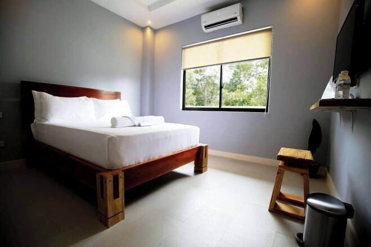 A bedroom with a bed, a window, a small stool, a trash bin, and a wall-mounted air conditioner.
