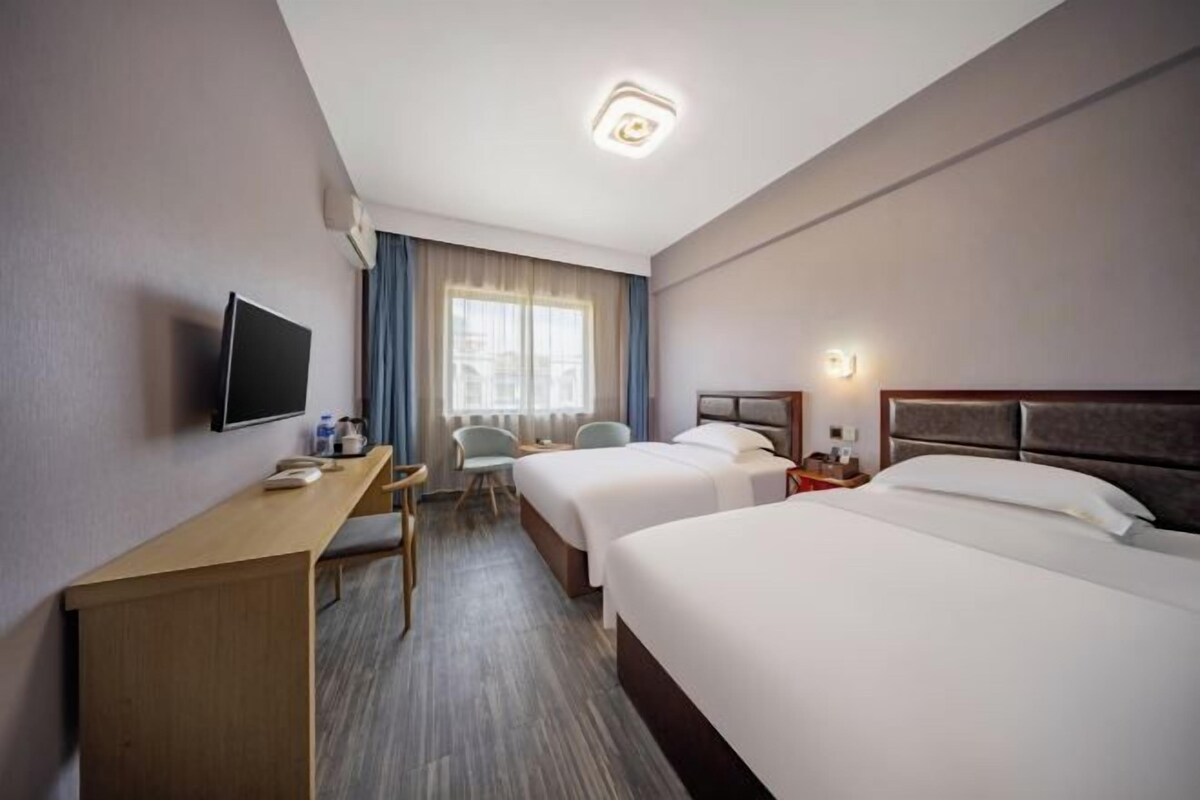 A hotel room with two beds, a desk, a TV, and a window with curtains.