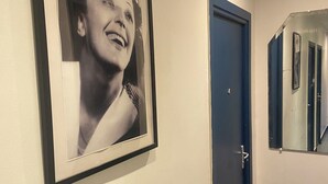 A hallway with a framed photograph on the wall, a closed blue door, and a mirror reflecting part of the hallway.