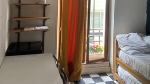 A room with a bed, a table, a shelf with plants, and a window with a curtain.