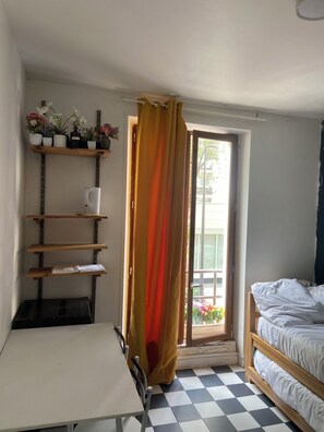 Ein Zimmer mit einem Bett, einem Tisch, einer Pflanzenregal und einem Fenster mit Vorhang.