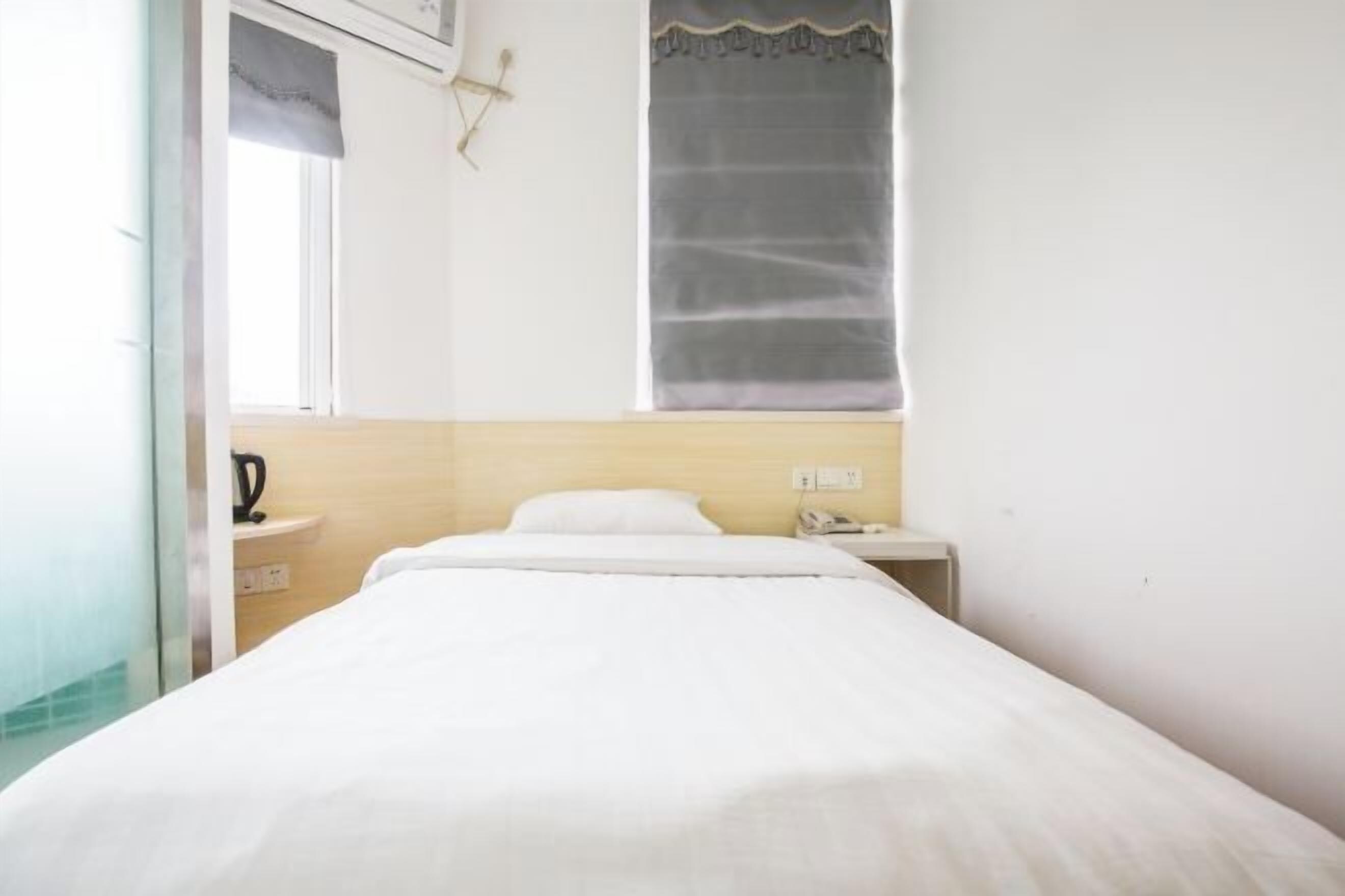 A single bed with white bedding, a bedside table with a phone, a window with a curtain, and a wall-mounted light fixture.