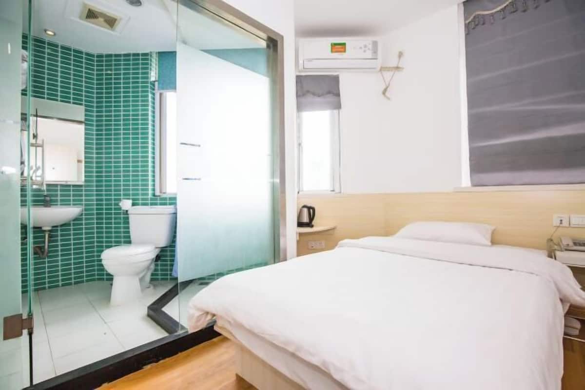 Quarto de hotel com divisória de vidro separando o banheiro, uma cama com roupa de cama branca, mesa de cabeceira com abajur e ar-condicionado fixado na parede.