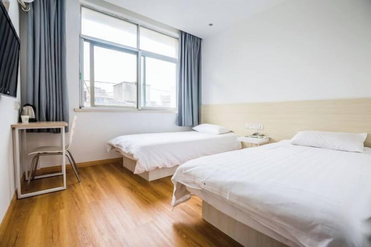 Quarto de hotel com duas camas de solteiro, uma escrivaninha com cadeira, mesa de cabeceira e janela com cortinas.