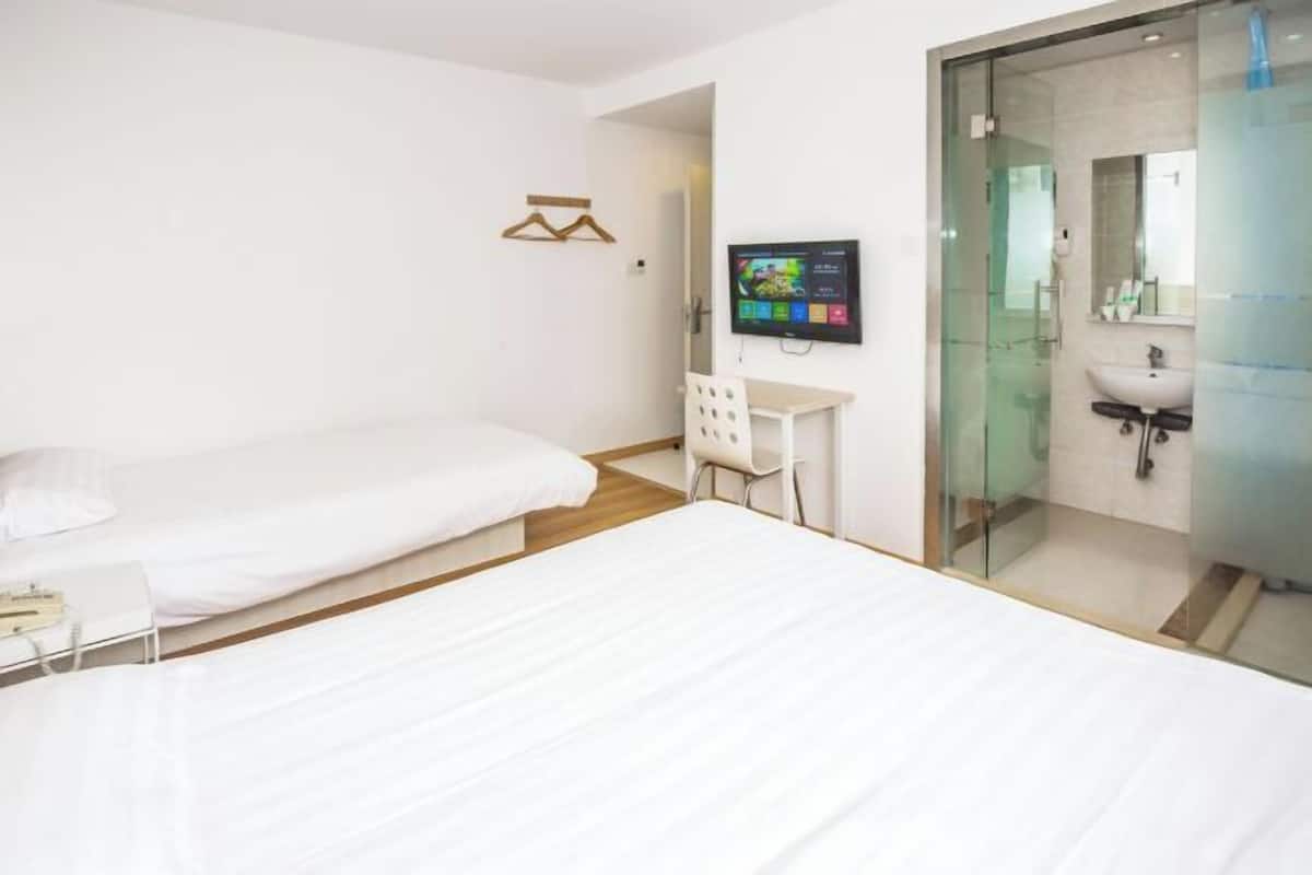 Quarto de hotel com duas camas, uma escrivaninha, televisão e chuveiro em box de vidro.