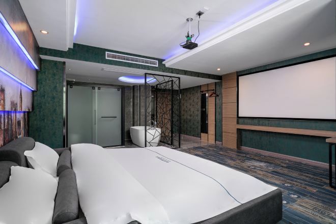 A modern hotel room with a large bed, a projector screen, and a seating area with a sofa and chairs.