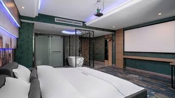 A modern hotel room with a large bed, a projector screen, and a seating area with a sofa and chairs.