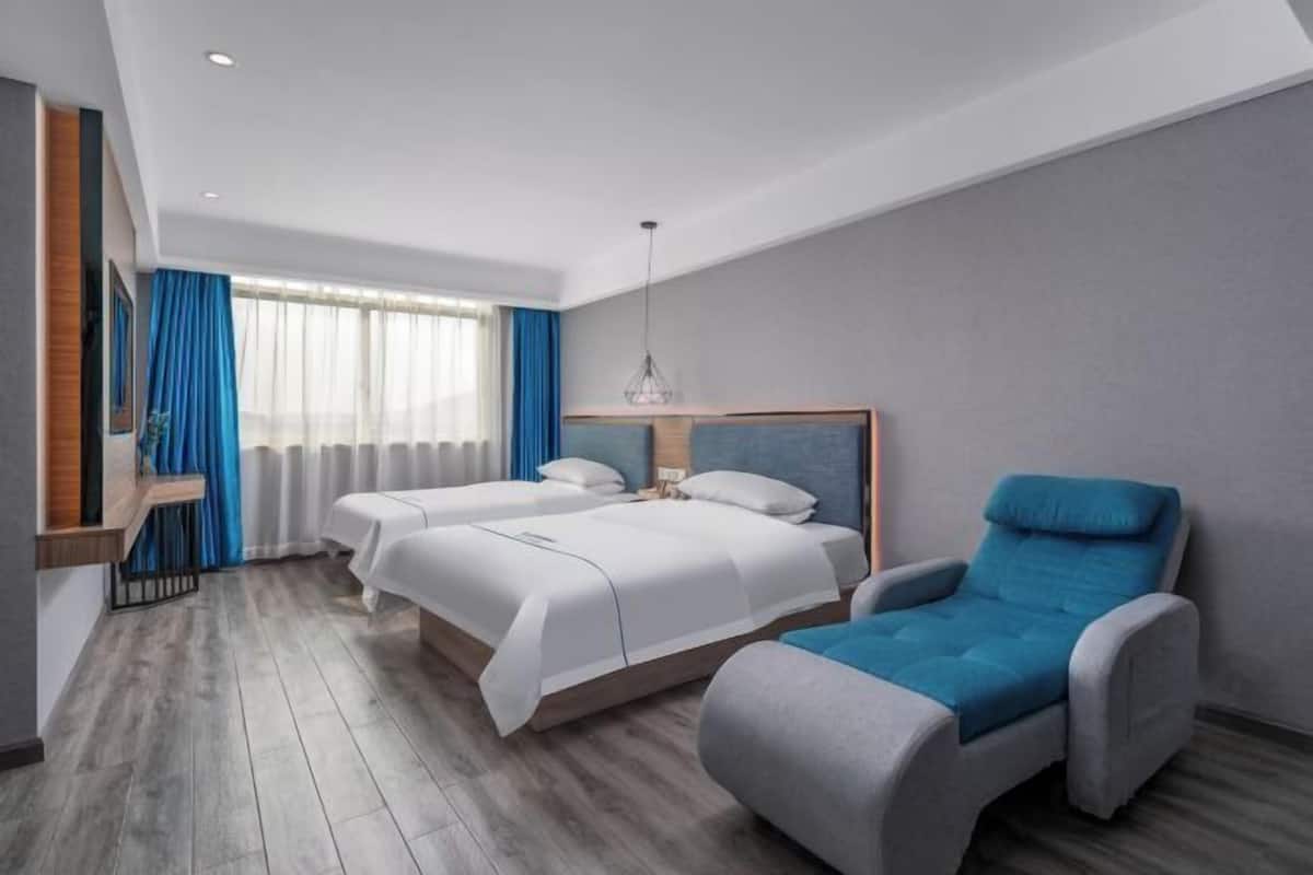Ein Hotelzimmer mit zwei Betten, einem blauen Sessel, einer Pendelleuchte und einem Fenster mit Vorhängen.