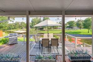 Een overdekt terras buiten met een barbecue, zitplaatsen en uitzicht op een meer en park.