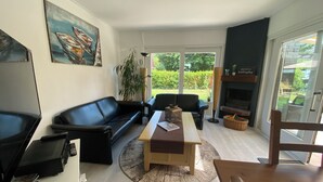 A modern living room with a black leather sofa, a wooden coffee table, and a TV on a stand. There is a large window with a view of greenery outside. - Heavenly vacation home near the sea (Bruinisse)