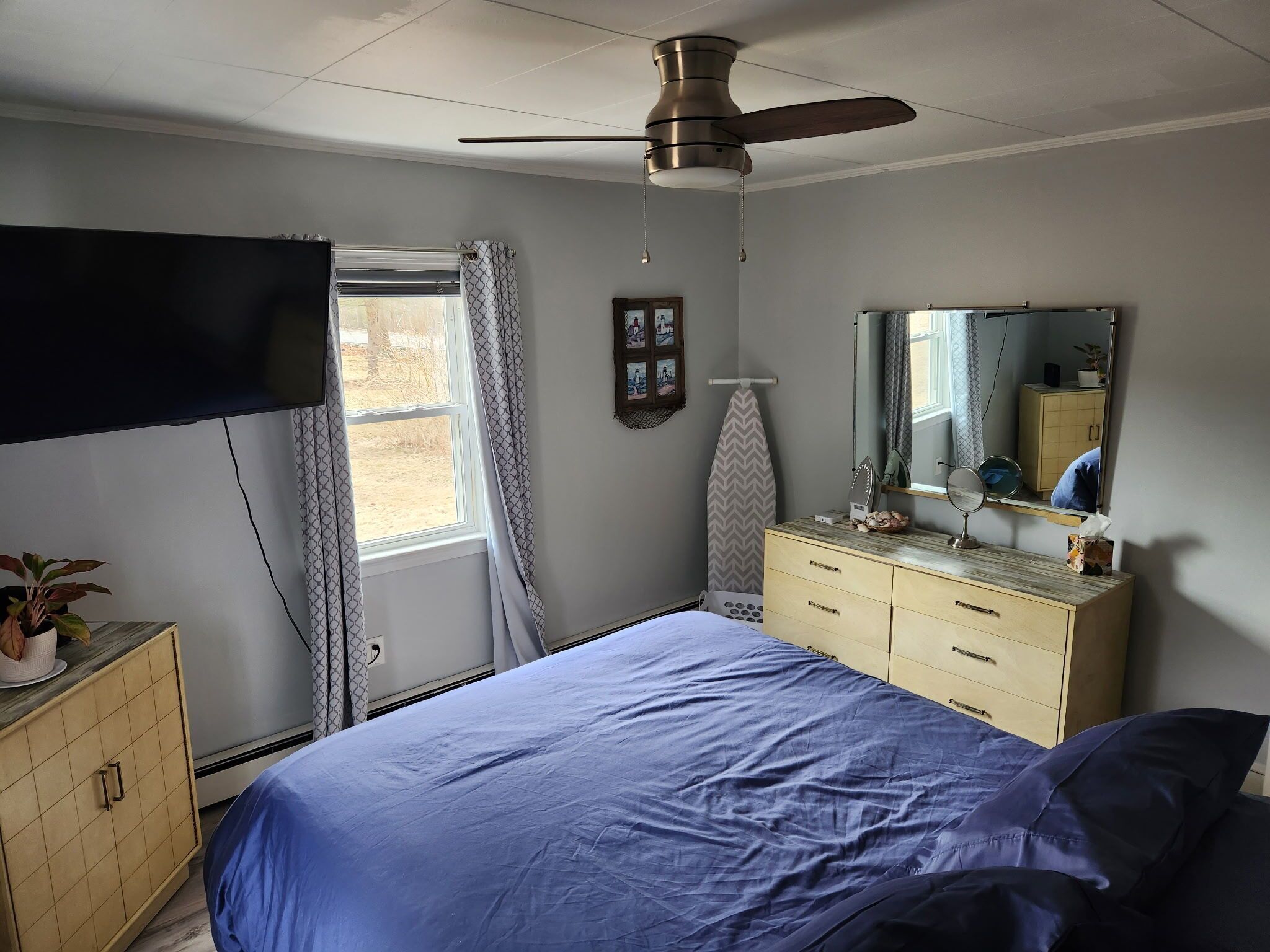 A bedroom with a bed, dresser, mirror, ceiling fan, and a window with curtains.