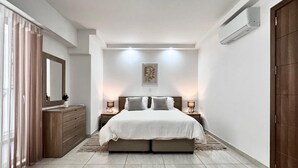 A bedroom with a bed, bedside tables, a dresser, a mirror, an air conditioning unit, and a door.