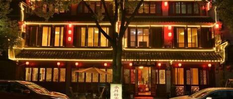 A traditional building with multiple floors, illuminated by warm lighting, and two cars parked in front.
