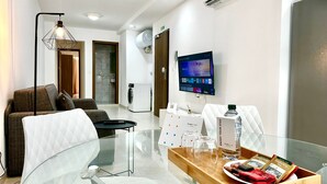 Un salon moderne avec une table basse en verre, un canapé, un téléviseur et un coin cuisine visible au fond.