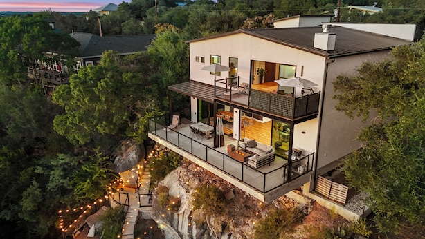 Une maison moderne à deux étages, dotée d’un balcon, d’un espace de détente extérieur et offrant une vue sur la forêt environnante.