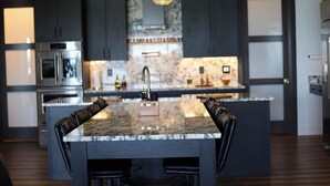 Une cuisine moderne avec des meubles de couleur sombre, un îlot central doté d’un plan de travail en marbre et un éclairage suspendu.
