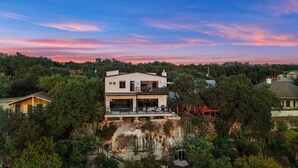 Une maison moderne à deux étages, dotée d’un balcon, entourée d’une végétation luxuriante et bordée d’une falaise.