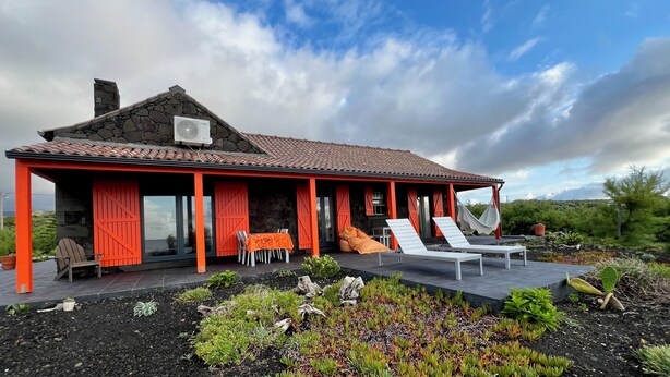 Een stenen huis met een rood dak en oranje luiken, met een terras met lounge stoelen en een hangmat.