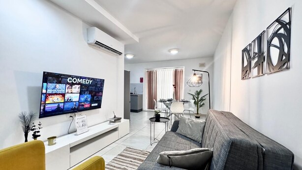 A modern living room with a flat-screen TV, a sofa, a yellow chair, and a dining area in the background.