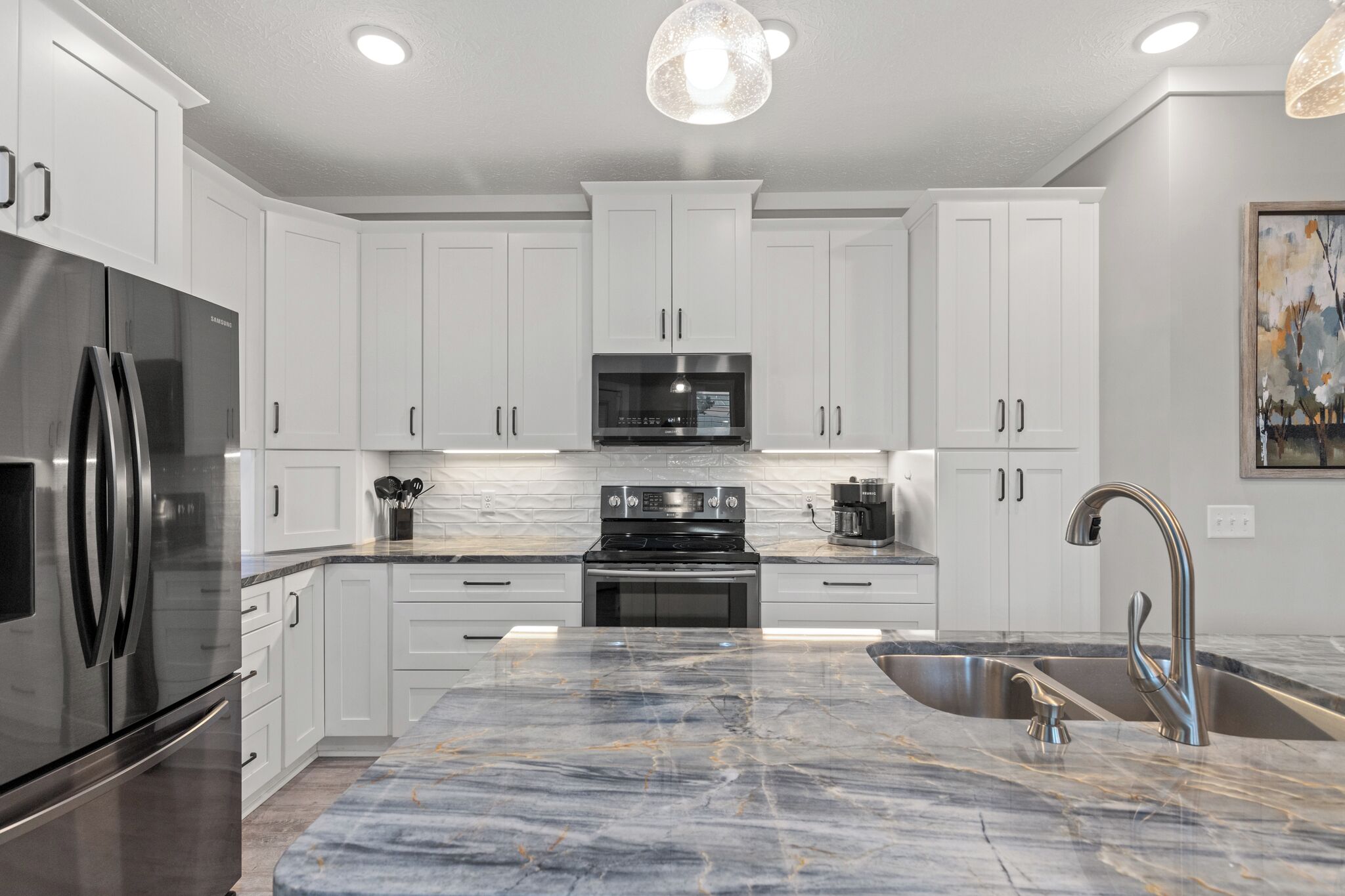 A modern kitchen with white cabinets, stainless steel appliances, a marble countertop, and a pendant light.