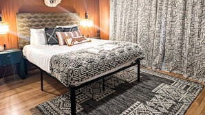 Een keurig opgemaakt slaapkamer met een bed, nachtlampjes, een kamerbreed tapijt met dessin en een gordijn met geometrische patronen.