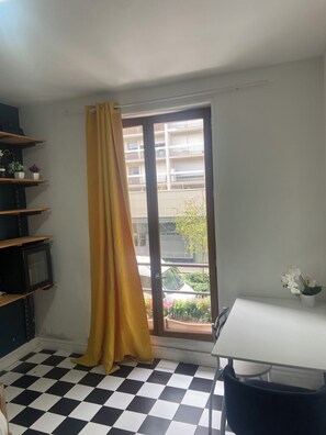Ein Zimmer mit Schachbrettmuster auf dem Boden, einem Fenster mit gelbem Vorhang, einem weißen Tisch und einem Stuhl.