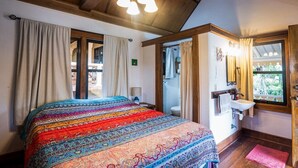 Un dormitorio con una cama grande, techo de madera, ventana con cortinas, baño con lavamanos y espejo, y una puerta que da a otra habitación.