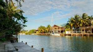 Un muelle de madera que conduce a una propiedad frente al agua, con un velero, palmeras y un edificio con balcón.