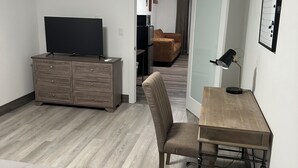 A compact hotel room with a bed, a desk, a chair, a television, a wardrobe, and a kitchenette in the background.