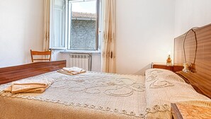 A bedroom with a bed, a chair, a bedside table with a lamp, a window with curtains, and a radiator.