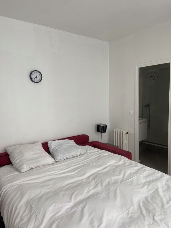 Une chambre à coucher comprenant un lit, une horloge murale, un radiateur et une porte donnant sur une salle de bain.