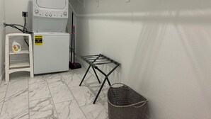 Un espacio para lavandería con lavadora, tendedero, cesto y estante con una botella de detergente.