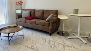 Un sofá de cuero marrón con una manta roja y dos almohadas, una mesita redonda con un recipiente verde, y una lámpara blanca sobre una mesa blanca.