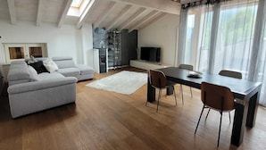 A spacious living area with a high ceiling, wooden beams, a grey sofa, a dining table with chairs, and a large window with sheer curtains. - Splendido Appartamento per il tuo Relax (Trentino-Alto Adige)