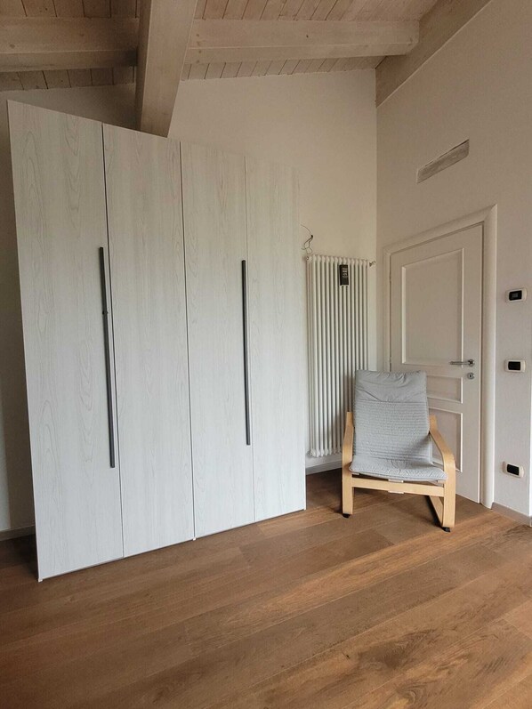 A room with a wooden floor, a white wardrobe with black handles, a radiator, and a light grey chair.