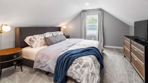Un dormitorio con cama, mesitas de noche, cómoda, televisor y ventana con cortinas.