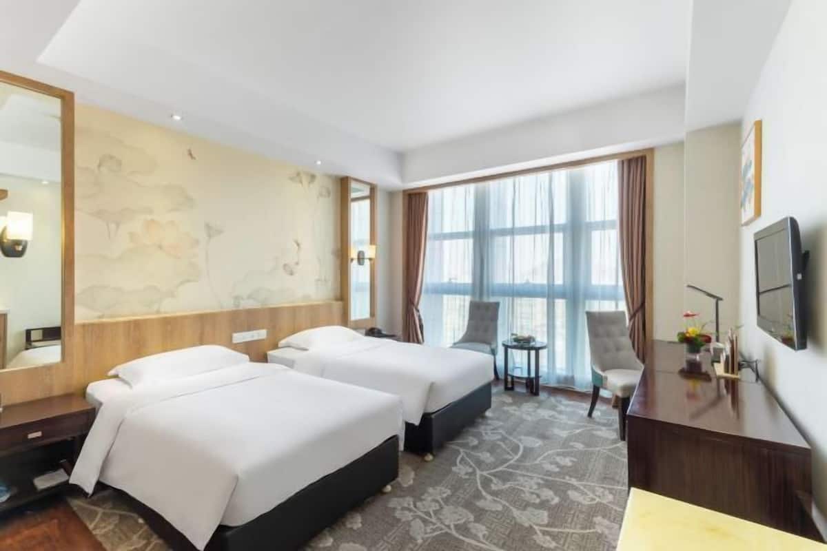 A hotel room with two beds, a desk, a chair, a TV, and a large window with curtains.