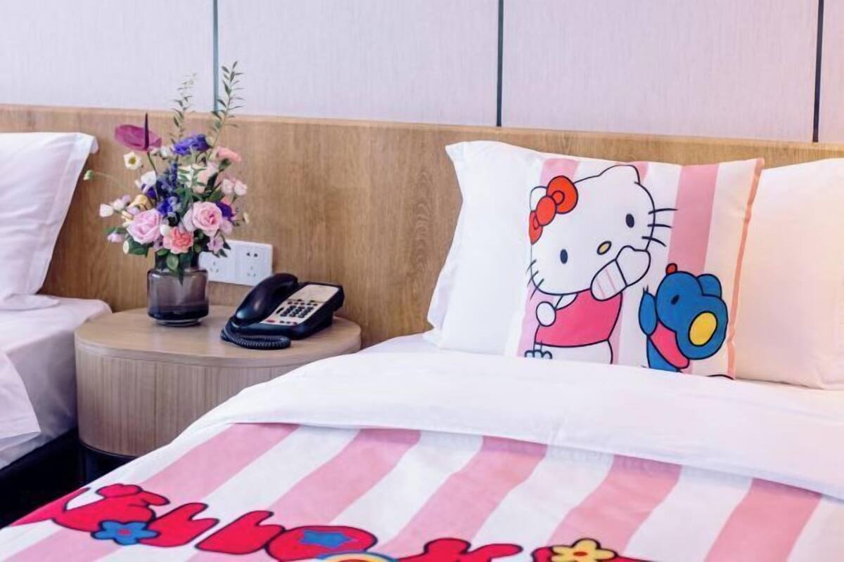 A hotel room with a bed featuring a Hello Kitty pillow, a bedside table with a phone and a vase of flowers, and a striped bedspread.