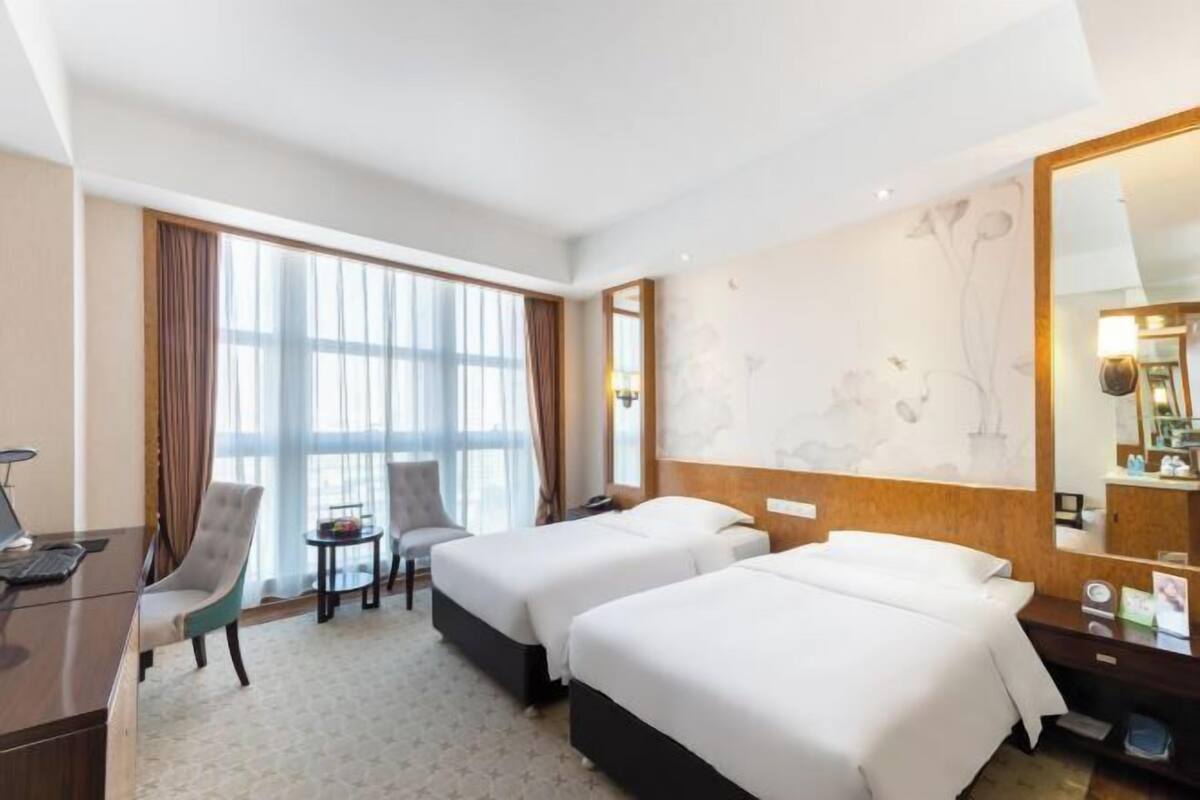 A hotel room with two beds, a desk, a chair, a small table, and a large window with curtains.