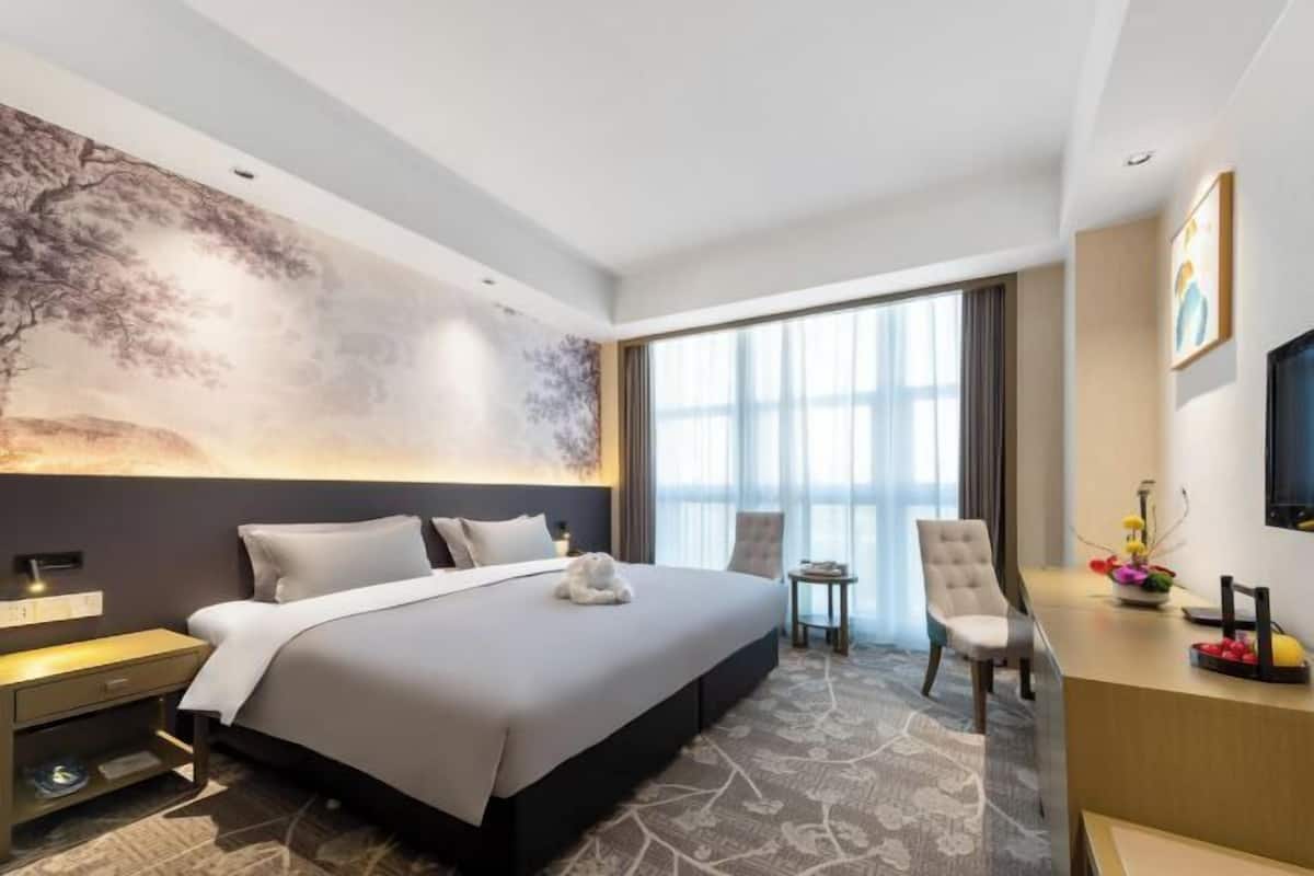A modern hotel room with a large bed, a desk, a chair, and a large window.