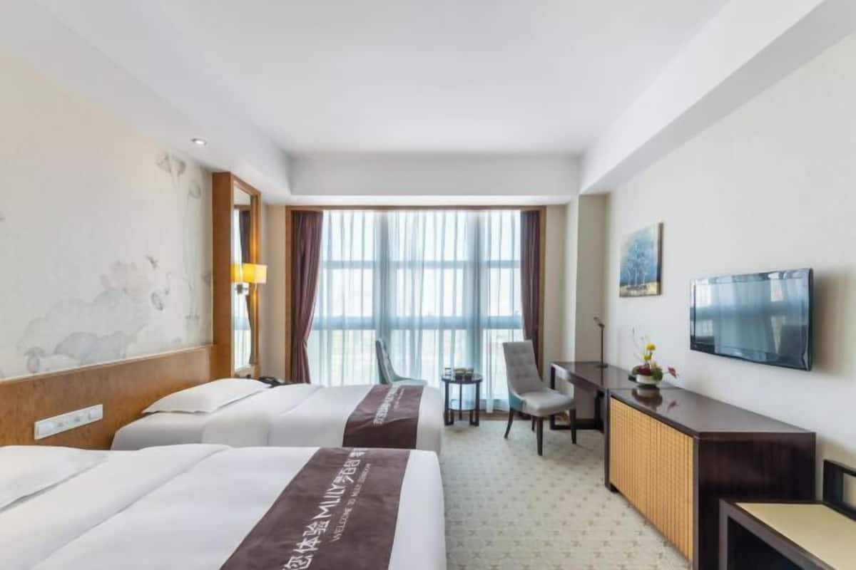 A hotel room with two beds, a desk, a chair, a TV, and a large window.