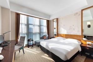 A hotel room with a large bed, a desk, a chair, a small table, and a large window with curtains.